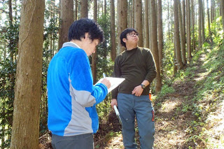 細い山道の林の中で一人がメモを取り、もう一人が立ち止まって木の上を見上げている森林調査中の写真