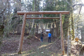 木製の鳥居の奥に木々に囲まれた小さな神社が見え、二人の男性が参道を歩いている後ろ姿の写真