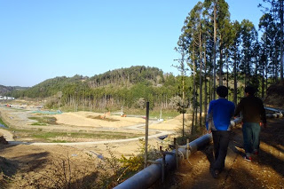 右側に細い山道とパイプが並び、左奥にある木々に囲まれた広い土地が見渡せる場所を歩く二人の後ろ姿の写真