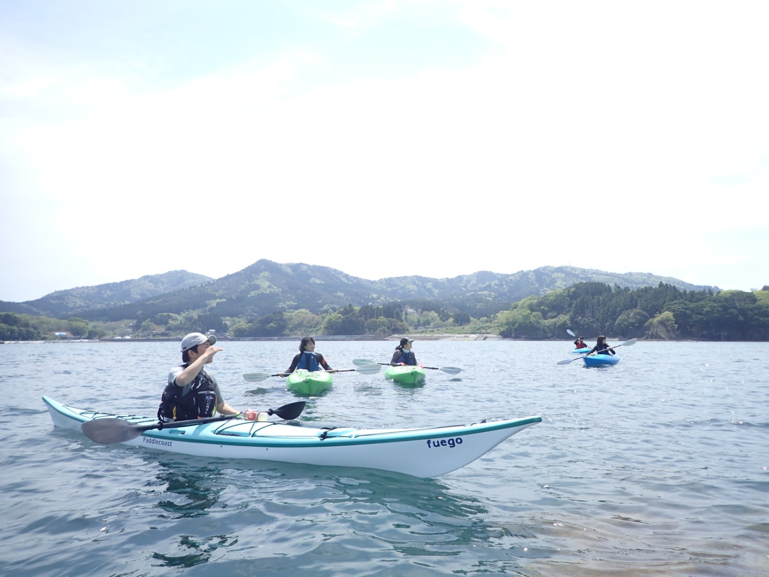 緑の山々を背景に、海の上で、白いカヤックに乗った人物がパドルを構えており、その奥に緑や青のカヤックに乗った他の人々がゆったりと浮かんでいる写真