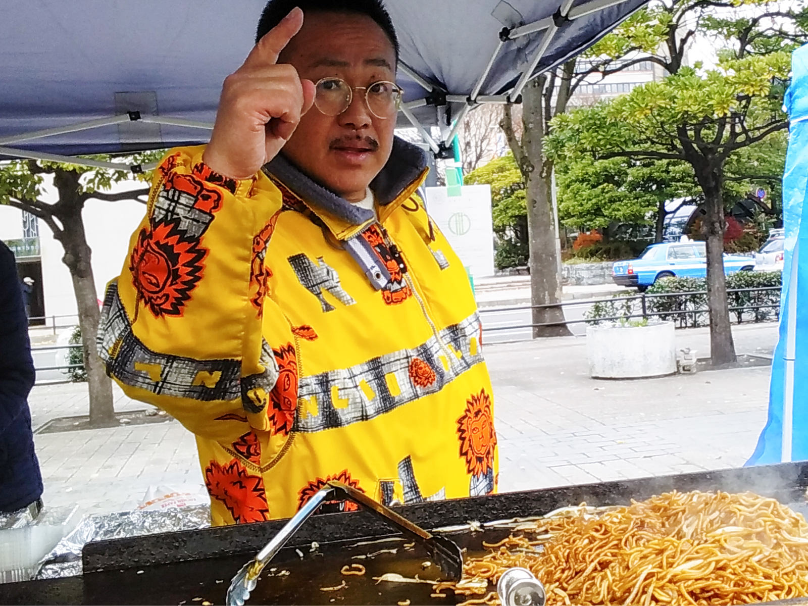 屋台の鉄板前で、黄色くて派手な柄の上着を着た男性が焼きそばを焼きながらカメラに向かって人差し指を立てている様子の写真