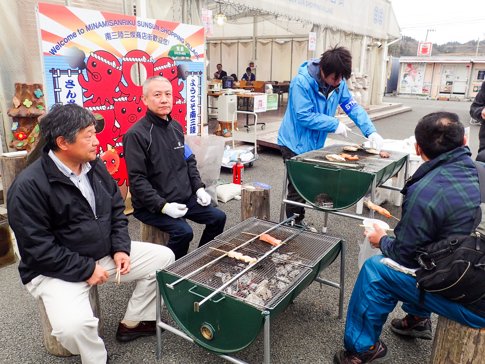 朝市の一角に設けられたコンロコーナーに2台の炭火焼の網が置かれ、串焼きや海産物などが焼かれている周りに数名の男性が座っている様子の写真