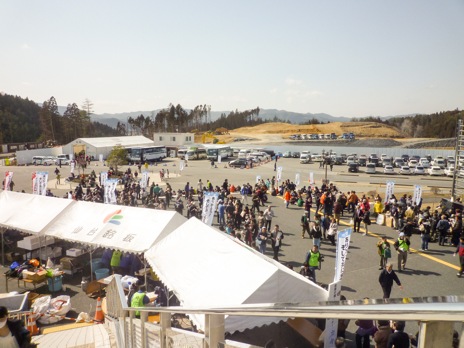 奥に山や森が広がるり、広い駐車場に多くの車が停まっている会場に白いテントが並び、大勢の人々でにぎわっている様子の写真