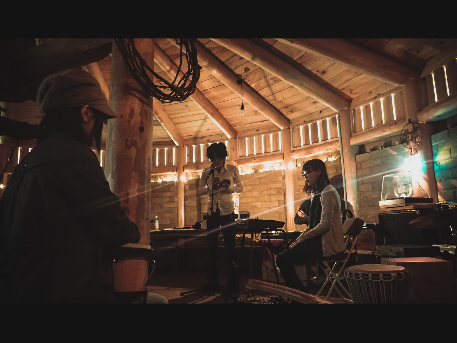 木造の室内で女性がキーボードの前に座り、白いシャツを着た男性がマイクの前に立っている様子を、太鼓を膝に乗せた女性の後ろ姿越しに撮影した写真
