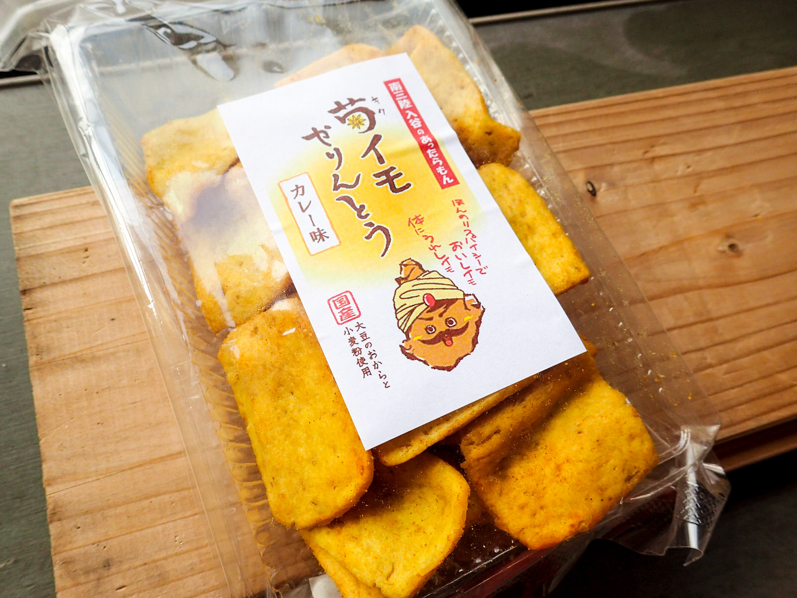 透明のパッケージの中に、四角形の黄色いかりんとうが入っており、「菊イモかりんとう カレー味」などの文字と頭に白いターバンを巻いた男性のイラストが載ったシールが貼られた商品の写真