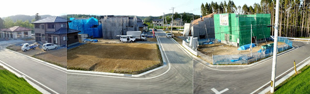 区画整備された住宅地に、完成した家や青や緑の工事用ネットに覆われた建設中の家々が並び、舗装された道路沿いや敷地内には車両が停待っている様子の写真