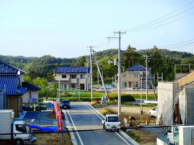 奥には緑の森の前に建つ、屋根に太陽光パネルを載せた2軒の住宅があり、その手前には複数の建設中の住宅があり、中央に写る舗装道路に沿って電柱が立ち並んでいる様子の写真