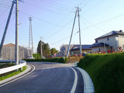 白いラインで縁取られ、カーブした道路が画面中央を通り、左側にはガードレール、道路の左右に複数の電柱が立ち、たくさんの電線が伸びており、奥には木々や住宅などの建物が見える写真