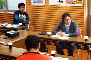 壁にポスターが貼られた会議室で、スーツ姿の男性や黒いシャツを着た男性が腕を組んで座り話を聞いている写真