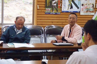 長机の一角でピンクのシャツを着た男性が、向かい側の席に座っている男性に向かって話しをし、隣の男性は机の上に置かれた資料に目を通しながら話を聞いている写真