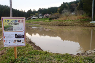 田んぼの脇に「メタン発酵液肥 実地試験中」と書かれた立て看板が立っており、その奥には水が張られた田んぼと山に囲まれた農村風景が広がる様子を写した田んぼの試験地の写真