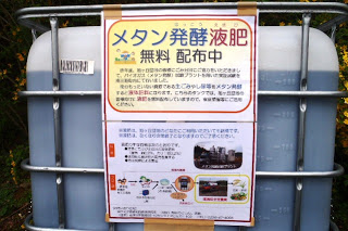 「メタン発酵液肥 無料配布中」と書かれた案内ポスターが大きな液体タンクの前面に貼られており、液肥の仕組みや使い方が図解付きで紹介されている様子を写した説明パネルの写真