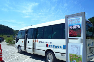 バス停の案内板が設置された砂利の駐車場に、ドアが開いた状態の白い送迎バスが停車している写真