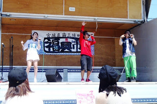 前の観客たちが見ているなか、屋外ステージで赤や白の服を着た3人が横一列に並び歌っているステージイベントの様子の写真