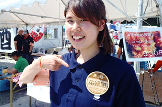 「応援団」と書かれた大きなバッジを胸につけた若い女性が、屋外のテントの前で笑顔で指さしている写真