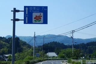 山と空が広がる背景に、右側にカーブする道路上に、赤い花の絵が描かれている「田束山 7キロメートル」と書かれた青い道路標識が立っている写真