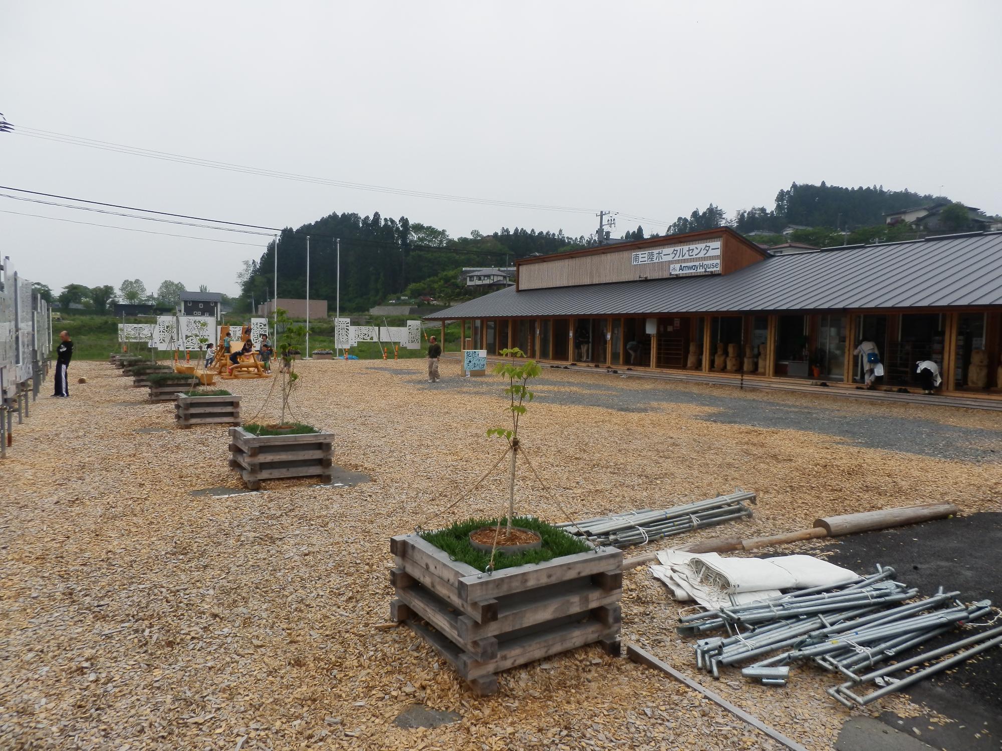 木製のプランターに植えられた木々が並んだ広場の奥に建つ木造の施設「南三陸ポータルセンター」の写真