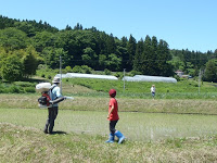 奥にはビニールハウスが見える田んぼのあぜ道で、赤い服の子どもと白い帽子の大人が話しをしている様子の写真