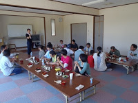 屋内の部屋で、複数の大人と子どもたちが長机を囲んで正座やあぐらで食事をしている写真