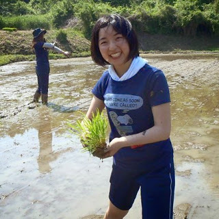青いシャツを着た女性が両手で苗を持ち、笑顔でカメラに向かって立っており、後ろには田んぼに入っているもう一人の人が見える田植え中の写真