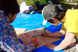 二人の人物がブルーシートの上でダンボールに「おら酒」とカラフルなペンで文字や絵を描いている看板作りの写真