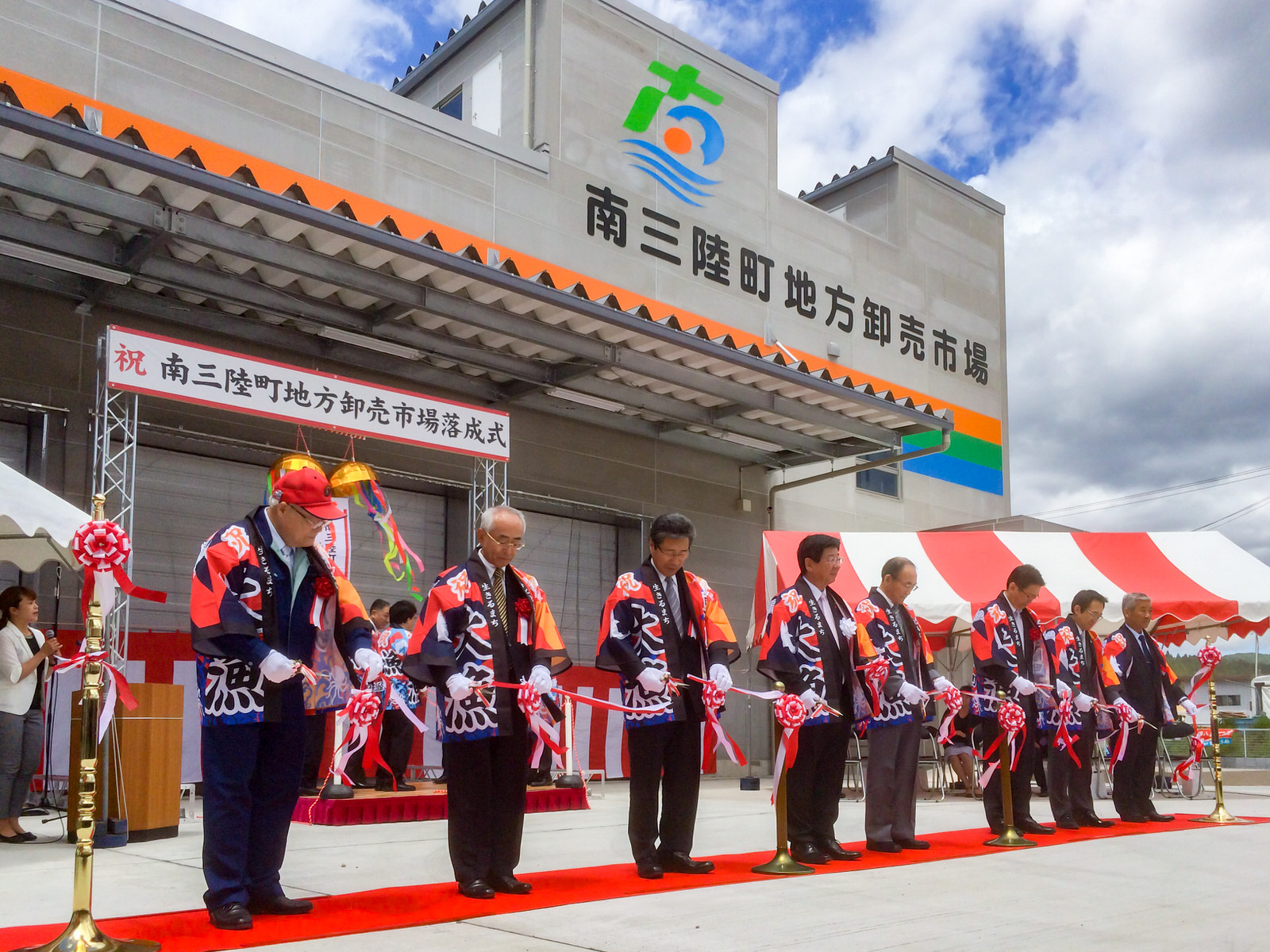 南三陸町のロゴマークと「南三陸町地方卸売市場」の文字が書かれた、大きな本設魚市場の正面入口前に設置された「祝 南三陸町地方卸売市場落成式」と書かれた横断幕の前に並んだ、大漁旗がデザインされた法被を着た関係者の男性たちがテープカットをしている様子の写真