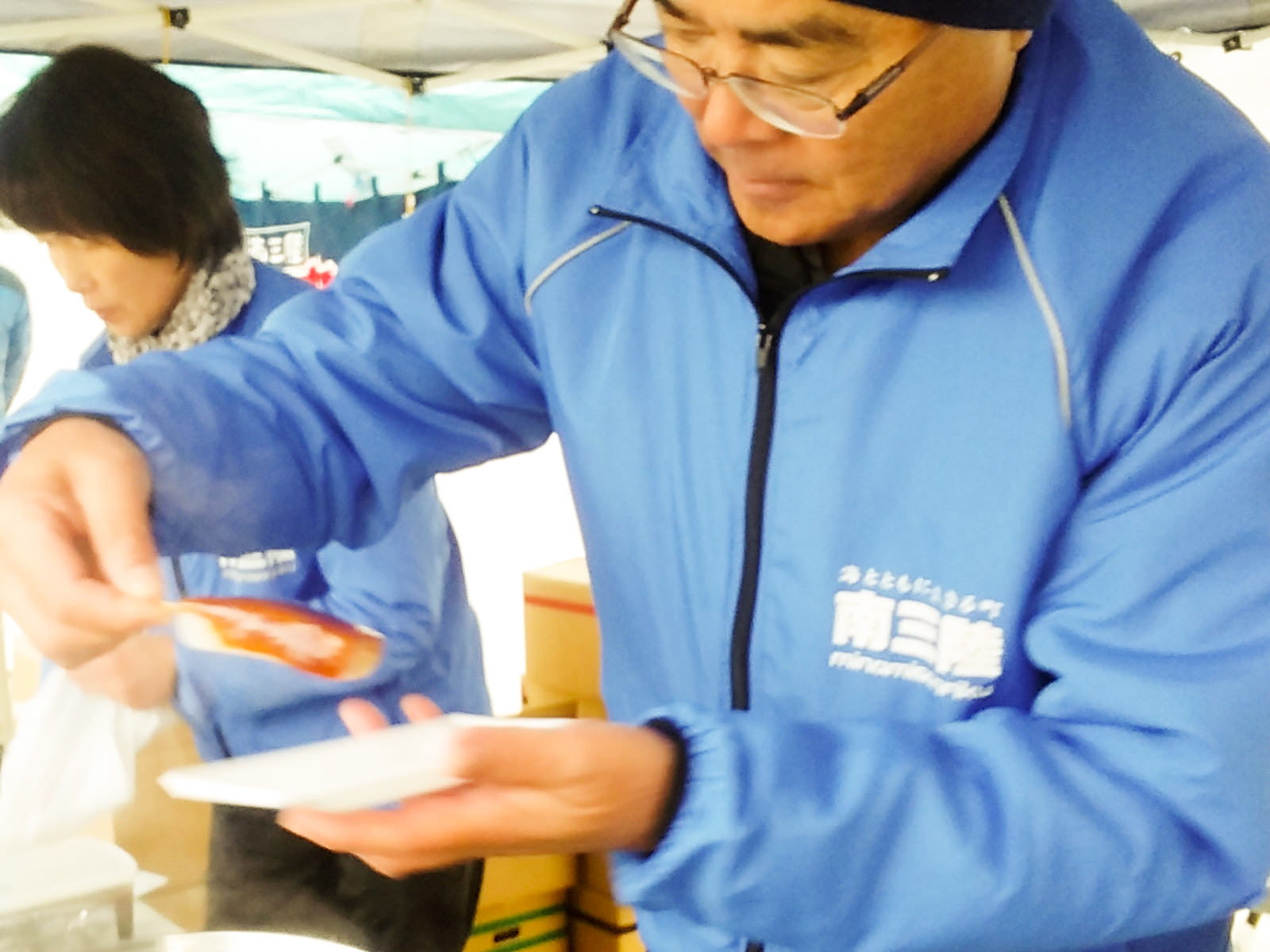 飲食ブースで、青いジャンパーを着た男性が味噌をたっぷり塗った串をさした食べ物を白い容器に盛りつけている様子の写真