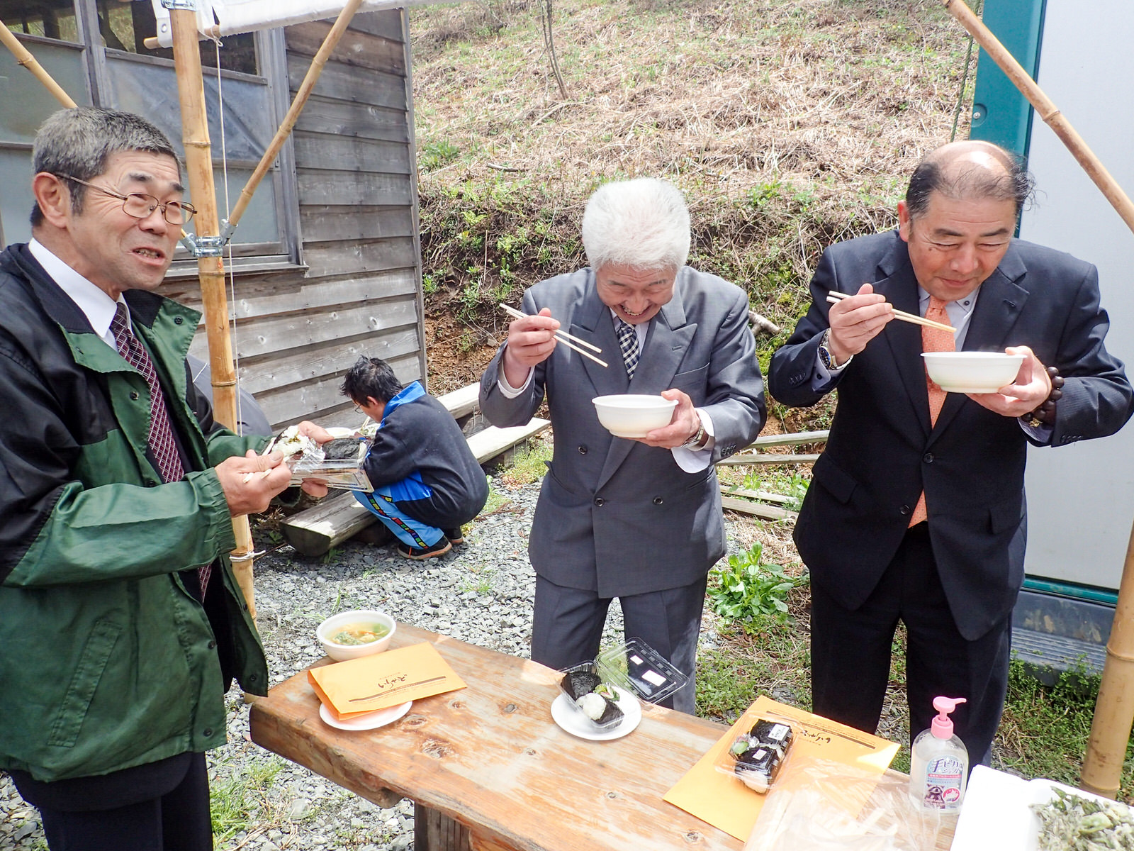 木のテーブルにパック入りのおにぎりなどが置かれ、その周りで3人の男性がお椀やおにぎりを持って笑顔で食事をしている写真