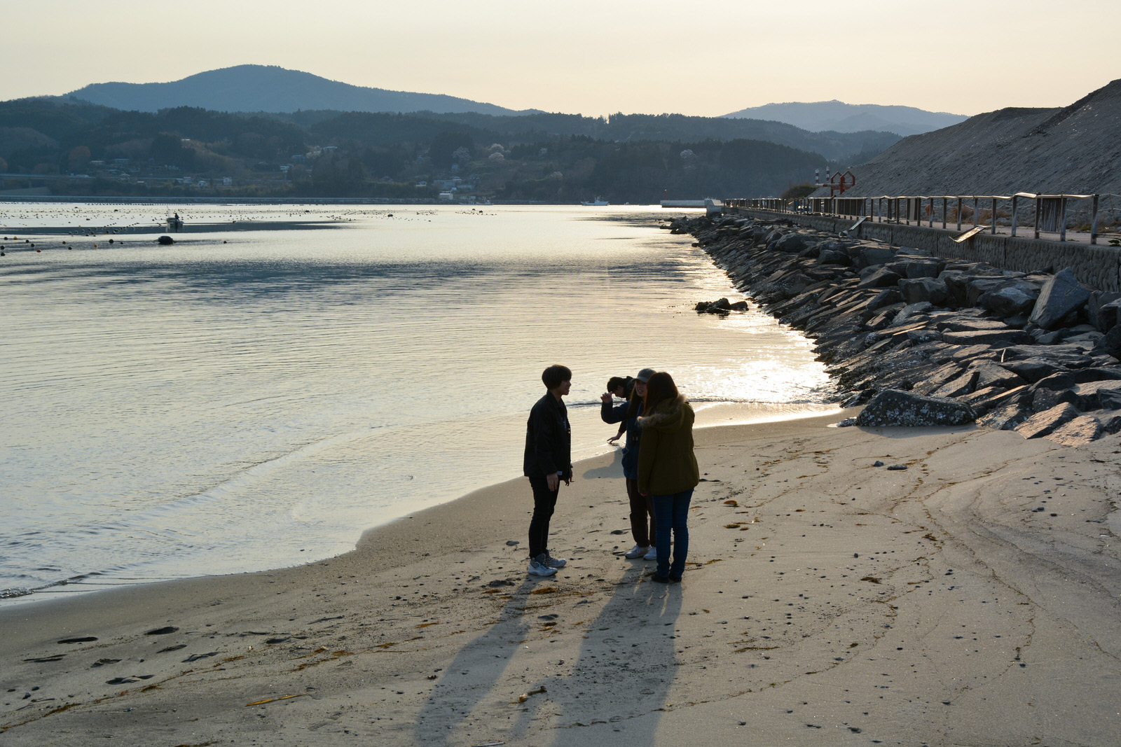 夕暮れ時の静かな海辺で、波打ち際で三人の若者が立ち話をしており、後ろには堤防と夕日に照らされた穏やかな水面が広がっている写真