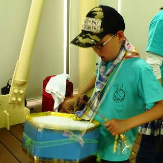 水色のTシャツを着た男の子が青い箱型の太鼓を首から下げて一生懸命叩いている写真