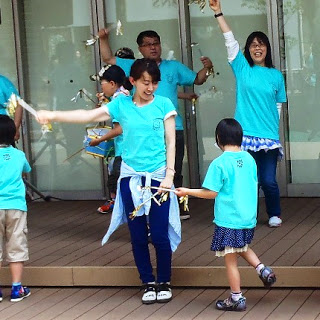 水色のTシャツを着た大人と子どもたちが屋外のステージ上で笑顔で手を振ったり踊ったりしている写真