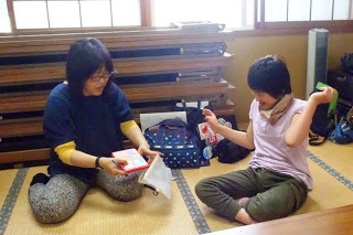 畳の部屋で床に座った紺色のシャツを着た女性がポーチから物を出し、向かい合ったピンク色のシャツを着た女性が喜んでいる写真