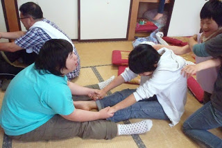 畳の部屋で複数の人がペアになり向かい合って座り、手をつないでストレッチのような運動をしている写真