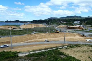 造成中の広い土地に重機やトラックが点在し、遠くに山々と海が見える青空の下で復興工事が進んでいる写真