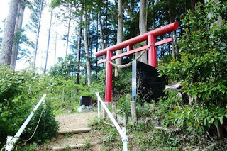 手前に白い手すりの付いた階段が続いており、黒い石碑が設置された森林の入口に赤い鳥居が建っている写真