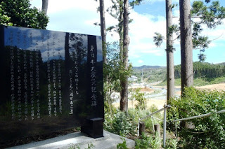 黒くて大きな石碑に白い文字で文章が刻まれており、背景には木々の隙間から造成地と空が見える写真