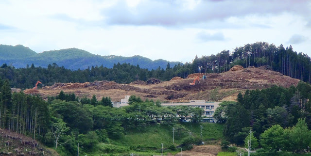 山の斜面に重機が入り木が大規模に伐採され、手前に学校のような建物がある開発中の風景の写真