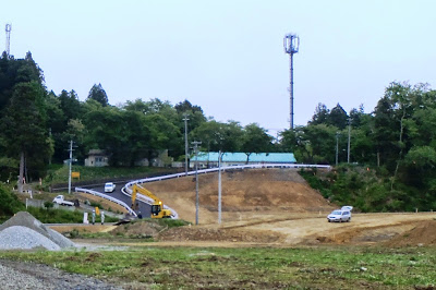道路が建設中の木々に囲まれた地域で、ショベルカーや車が行き交っている写真