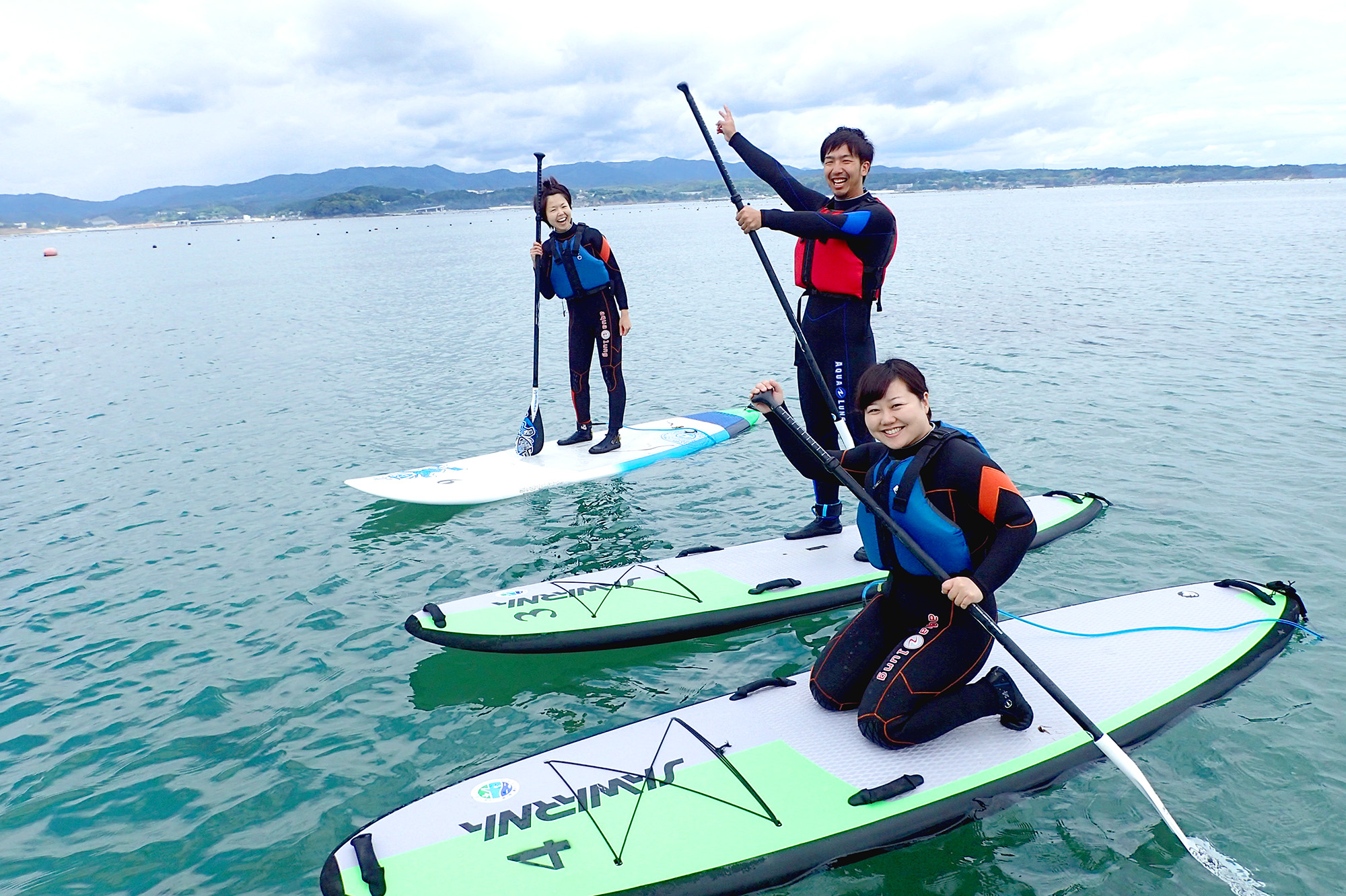 遠くに山並みが見える海の上で、3つ並んで浮かんだボードの上で膝をついて座っている女性リポーターとパドルを持って立っている男性、女性の写真
