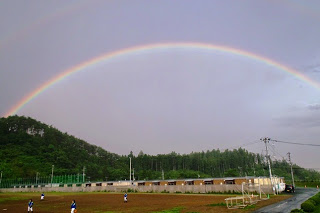 広いグラウンドとその奥に並ぶ建物の上空にくっきりとした虹が弧を描いてかかっている写真