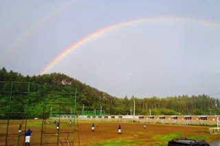 山と林を背景にしたグラウンドで野球の練習をしている中学生たちの頭上に大きく虹がかかっている写真