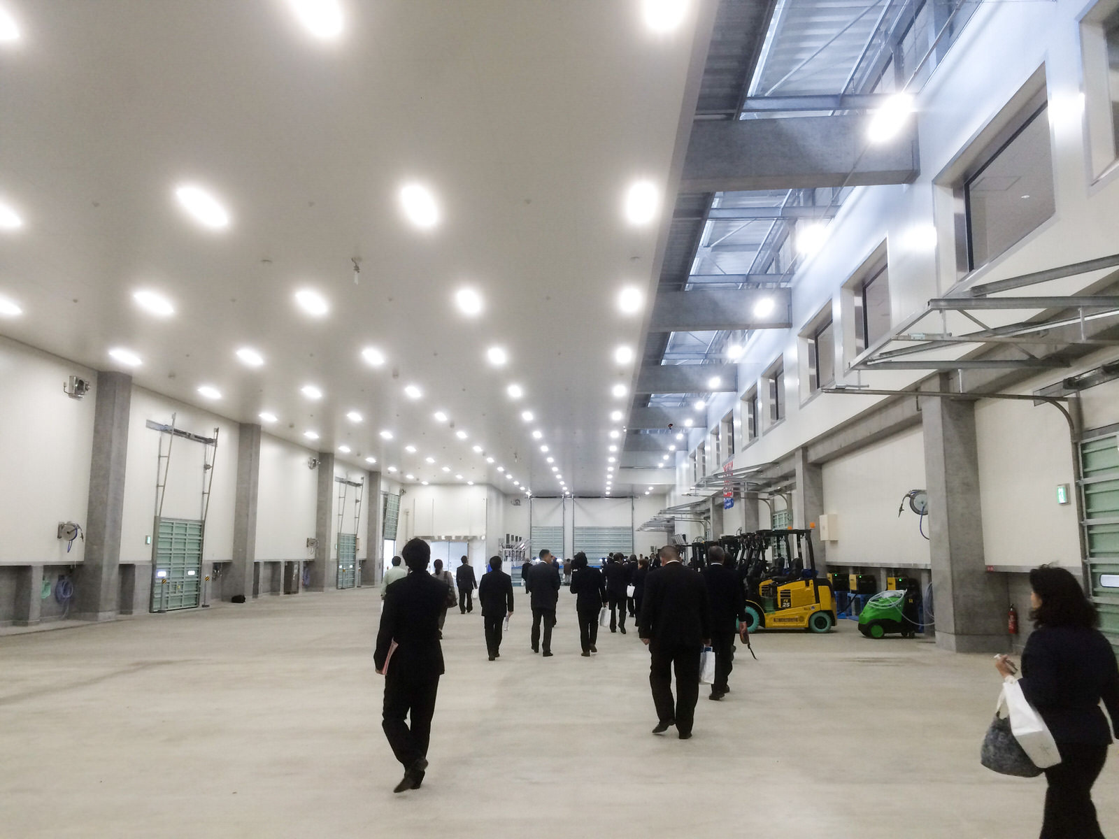 スーツ姿の大勢の人々が、明るい照明の広々としたコンクリート床で、右側にフォークリフトが数台並んでいる市場内を見学している写真