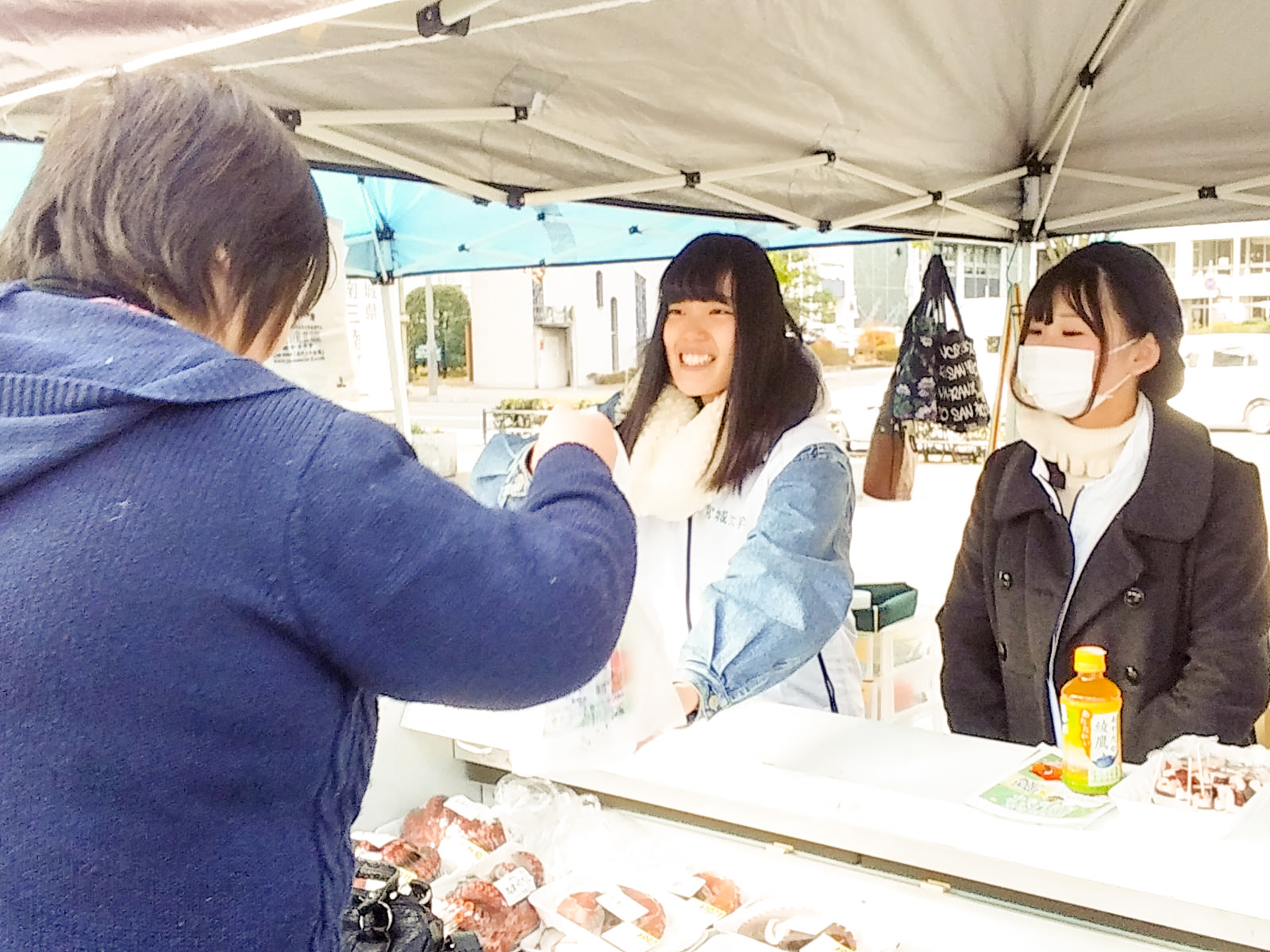 復興市の販売ブースで、青色の上着を着た客が笑顔の女性スタッフ二人と会話しながら商品を受け取っている様子の写真