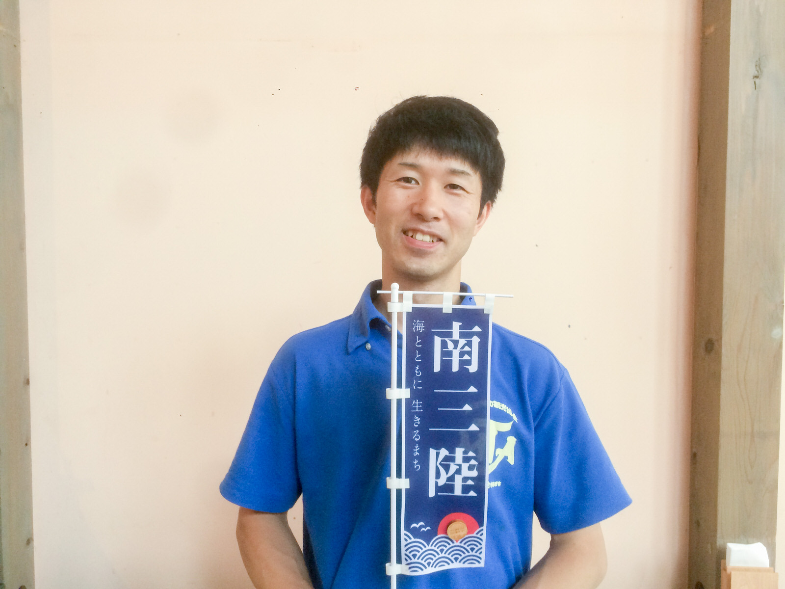青いポロシャツを着た男性が、「南三陸 海とともに生きるまち」と書かれた小さな青い旗を手に持って笑顔で立っている写真