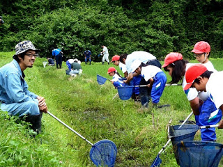 細い用水路の水面を網でかき回しながら探す子どもたちと、それを笑顔で見ている大人の男性が一緒に写っている写真