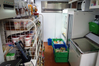 移動販売車の通路の両側に、カップ麺やお菓子、飲料などの棚や冷蔵庫が設置されており、手前にはレジと無線機が置かれている写真