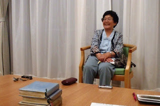 手前のテーブルには眼鏡ケースや録音機が置かれ、白いカーテンの前の椅子に年配の女性が笑顔で座っている写真