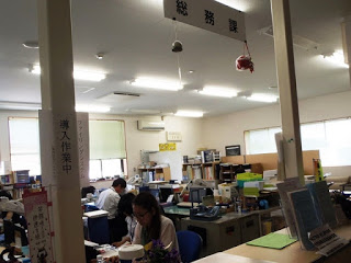 役場の総務課のカウンターの近くの席で机に向かっている亀山さんの写真