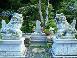 白い石造りの狛犬が左右に並び、その奥にある小さな石碑の横に花が供えられている写真