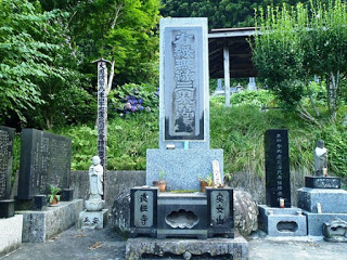 緑の草木の前に大きな石碑があり、左に小さなお地蔵様、右に新しい黒い石碑が立っている写真